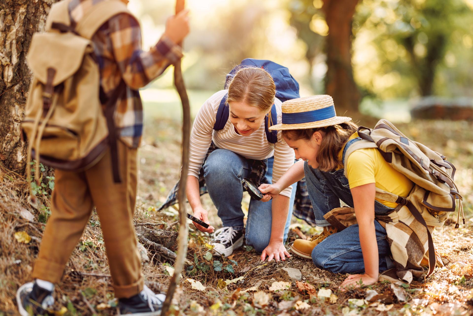 Happy mother and two   children in   with backpacks examining environment through magnifying glass in forest