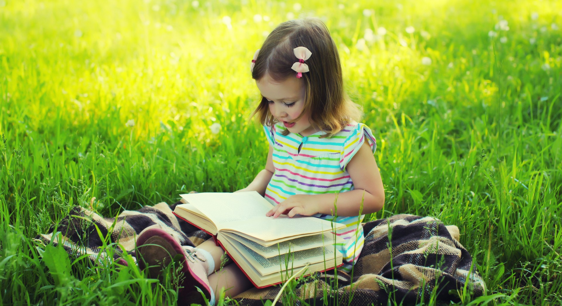 Happy little girl child reading book sitting on the grass in summer park